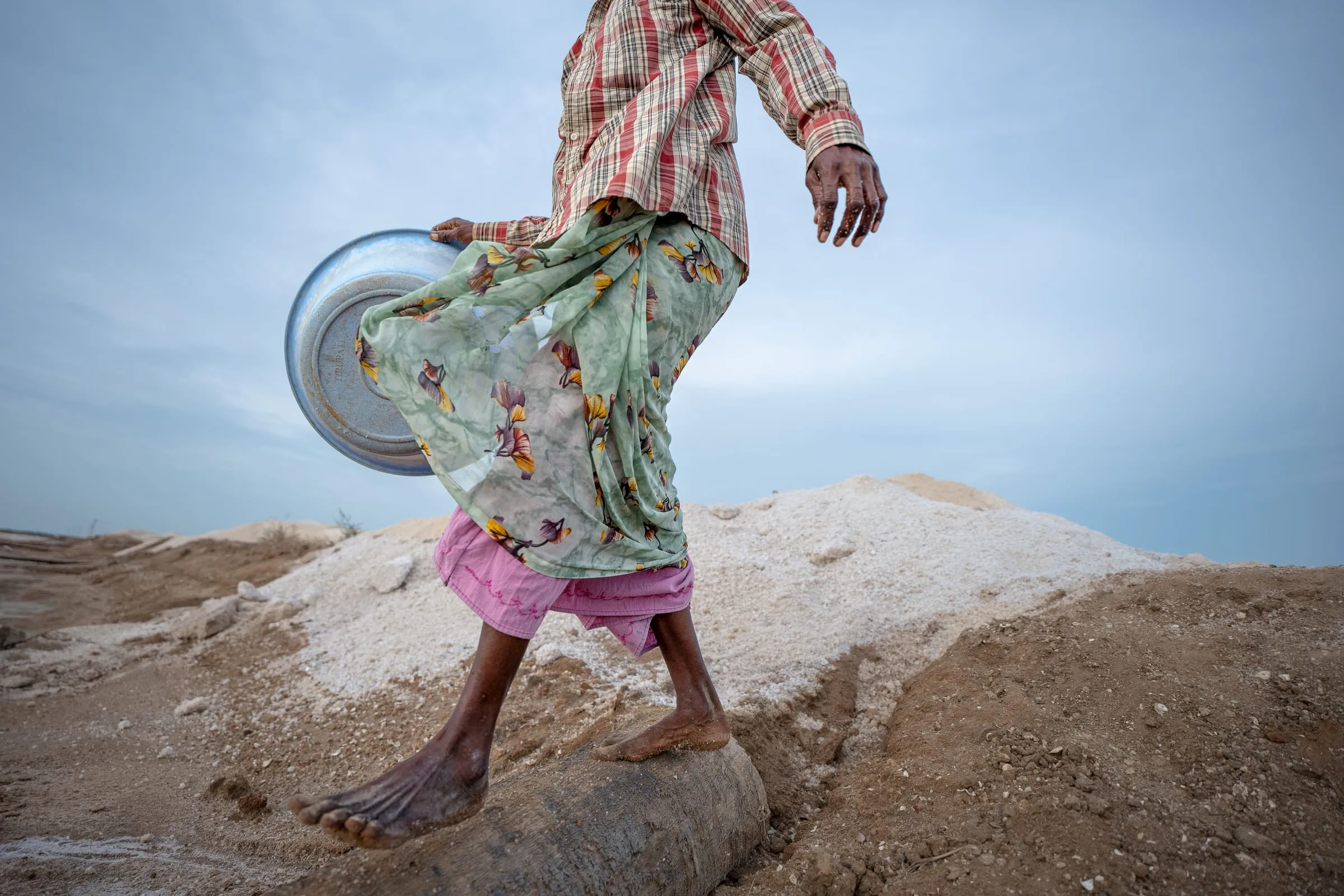 A woman carries salt in an aluminum satti (bowl) in Tamil Nadu, India. Women make up as much as 40% of the salt pan labor force, typically engaged in scraping, carrying, and drying salt. Loads can weigh up to 25 kilograms.
