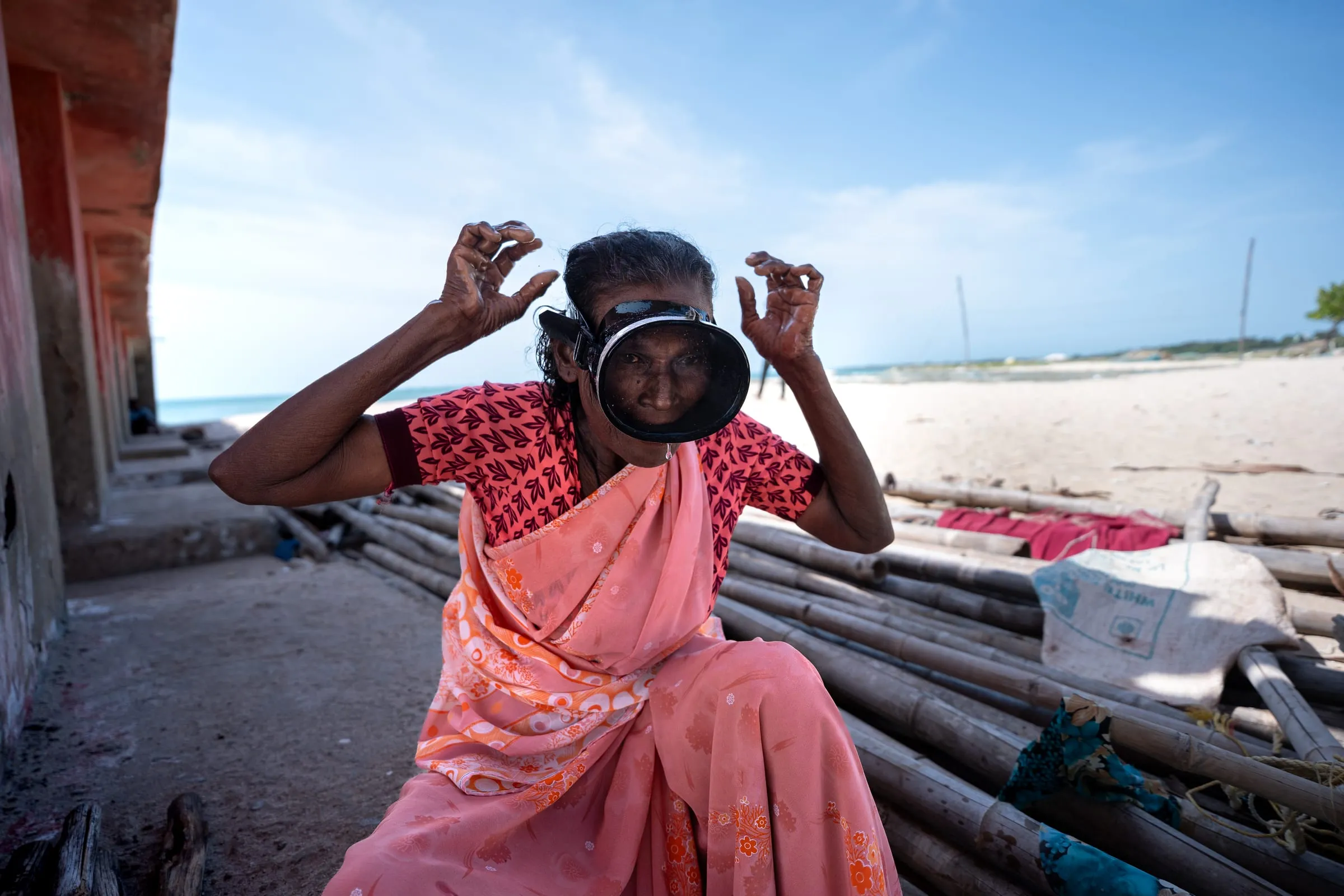 Mary, 57, poses with her diving mask outside her home in Rameswaram, on Pamban Island. Like hundreds of women divers, she works for hours underwater wearing a saree and using only basic gear. Protection is minimal, and sharp coral and rocks make each dive dangerous. The nearest medical facility is eight kilometers away, across the Pamban Bridge, leaving divers with little access to healthcare.